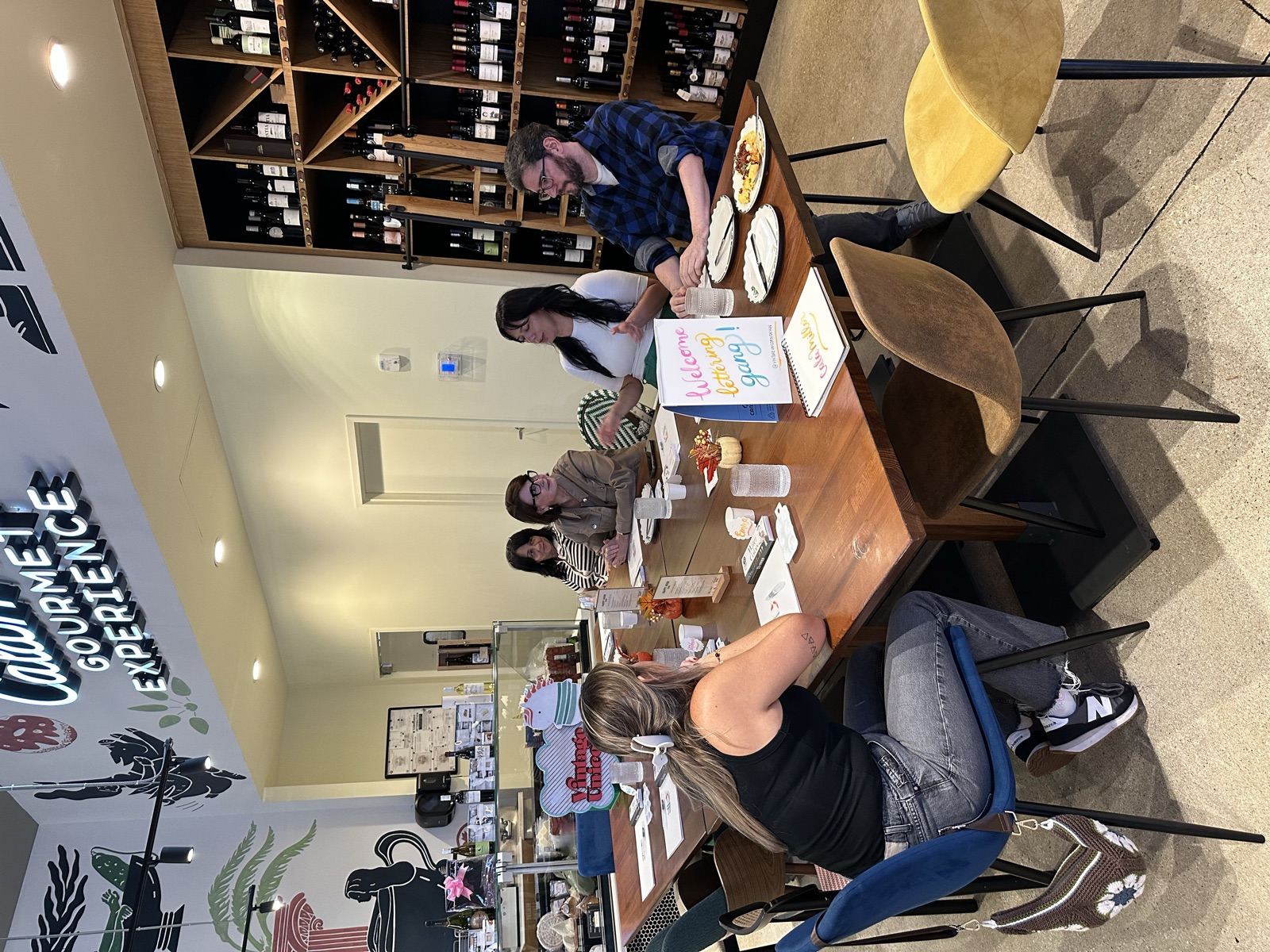 Brush lettering class seated at a long wooden table inside CalaMillor Gourmet Experience in Miami, with wine shelves in the background
