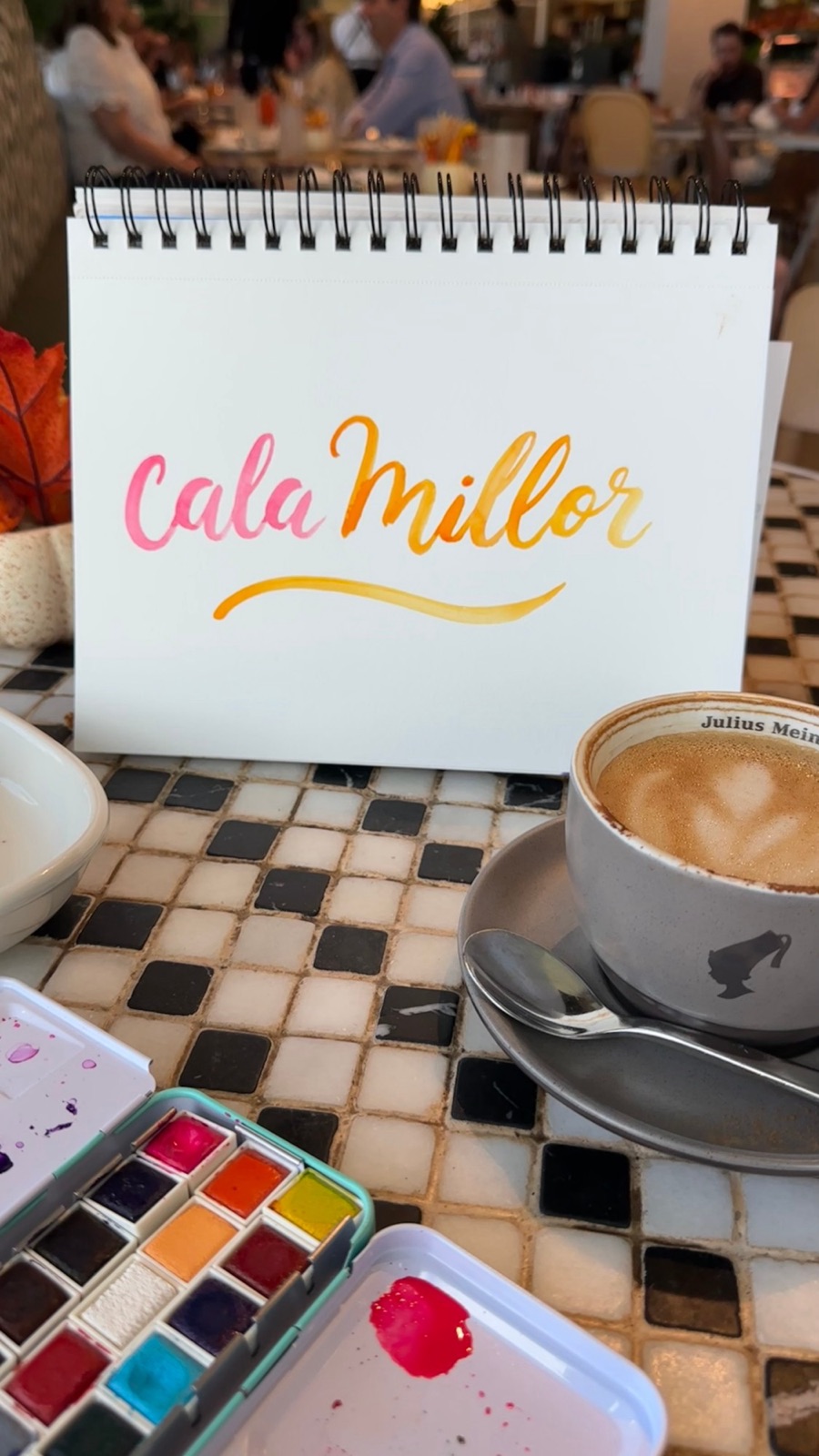 Cala Millor hand-lettered in pink and orange watercolor brush lettering on a sketchbook next to a latte and a watercolor palette